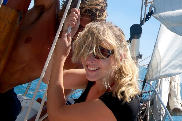 Two people on a sailboat smiling, holding onto ropes under a clear blue sky.