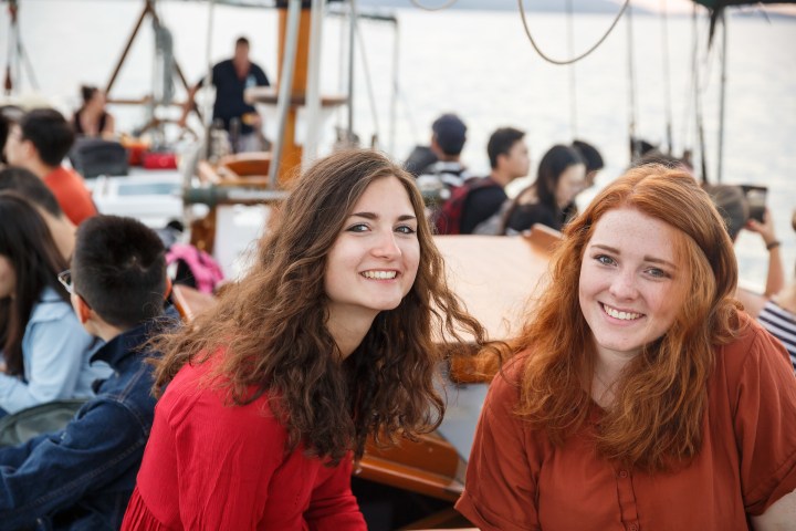 Two women smiling on a crowded boat with people in the background.