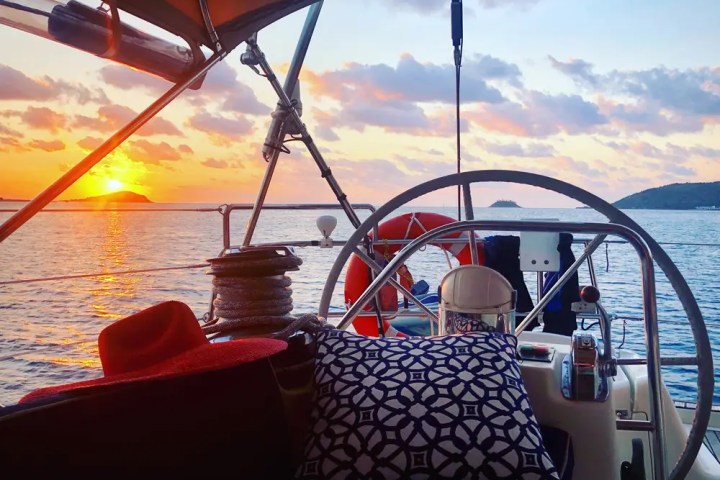 Sailboat cockpit at sunset, view of sea and distant islands, with cushions and a red hat.