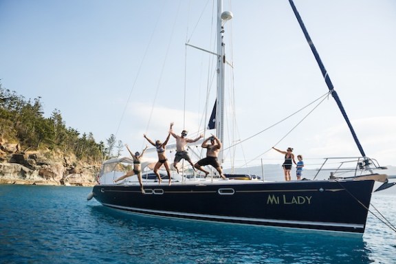 People joyfully posing on a sailboat named 'Mi Lady' in clear blue water near a rocky shore.