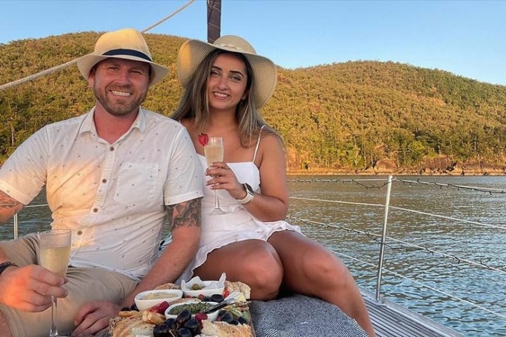 Couple on a boat with drinks and snacks, wearing sun hats, with a scenic backdrop.