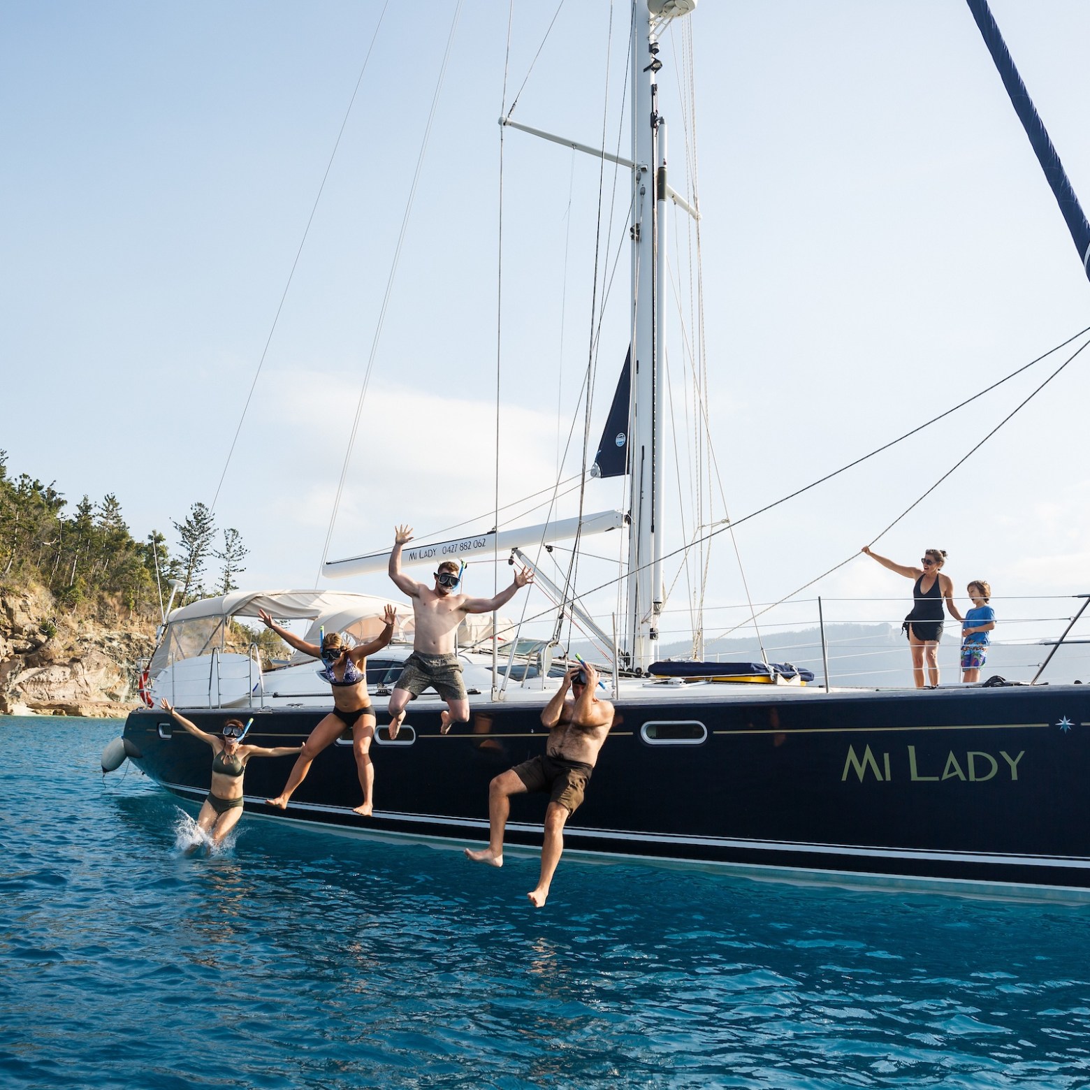 People jumping off a sailboat named 'Mi Lady' into the ocean near a rocky shore.
