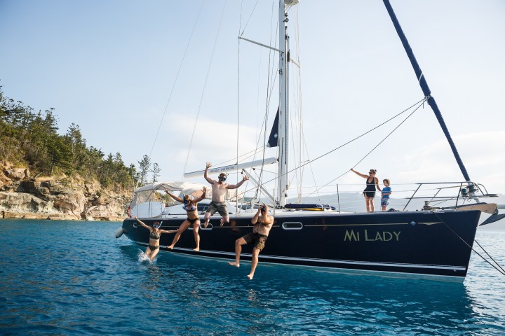 People jumping off a sailboat named 'Mi Lady' into the ocean near a rocky shore.